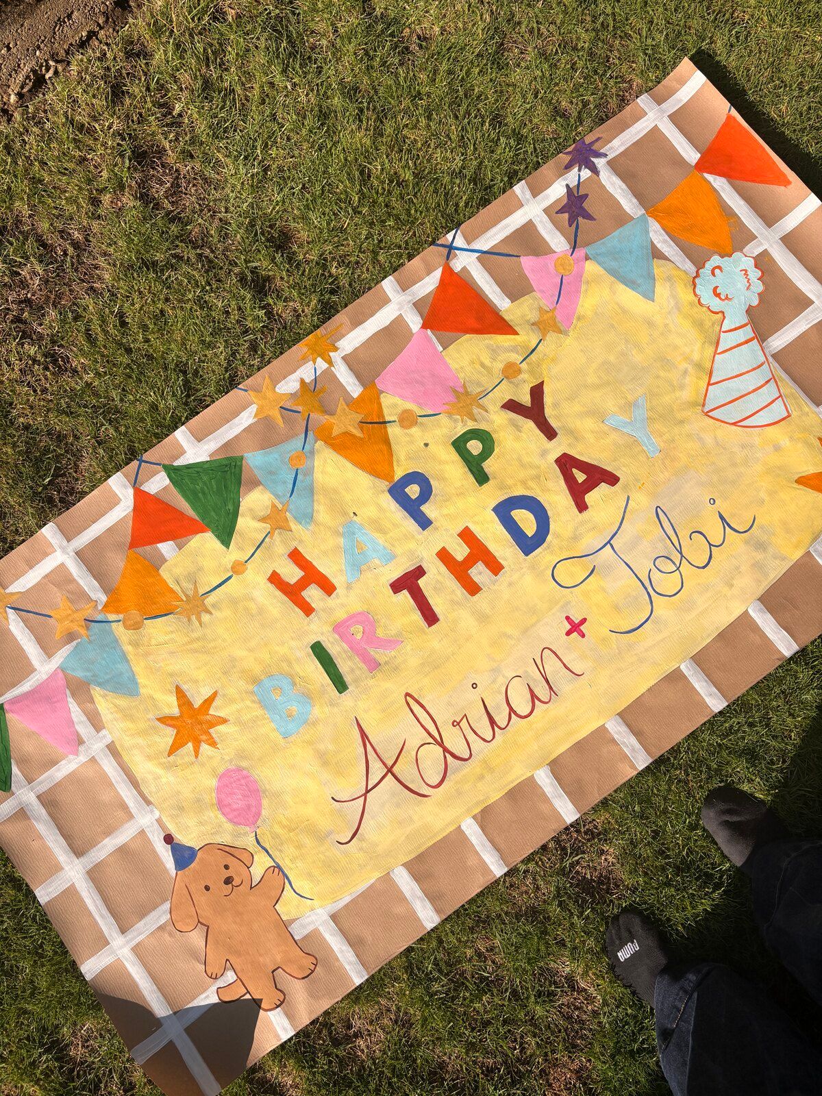 A kraft-paper banner with a white grid pattern and a pale yellow center, strung with colorful bunting and stars. It reads “Happy Birthday” in multicolored letters and “Adrian + Tobi” in script, with a puppy holding a balloon.