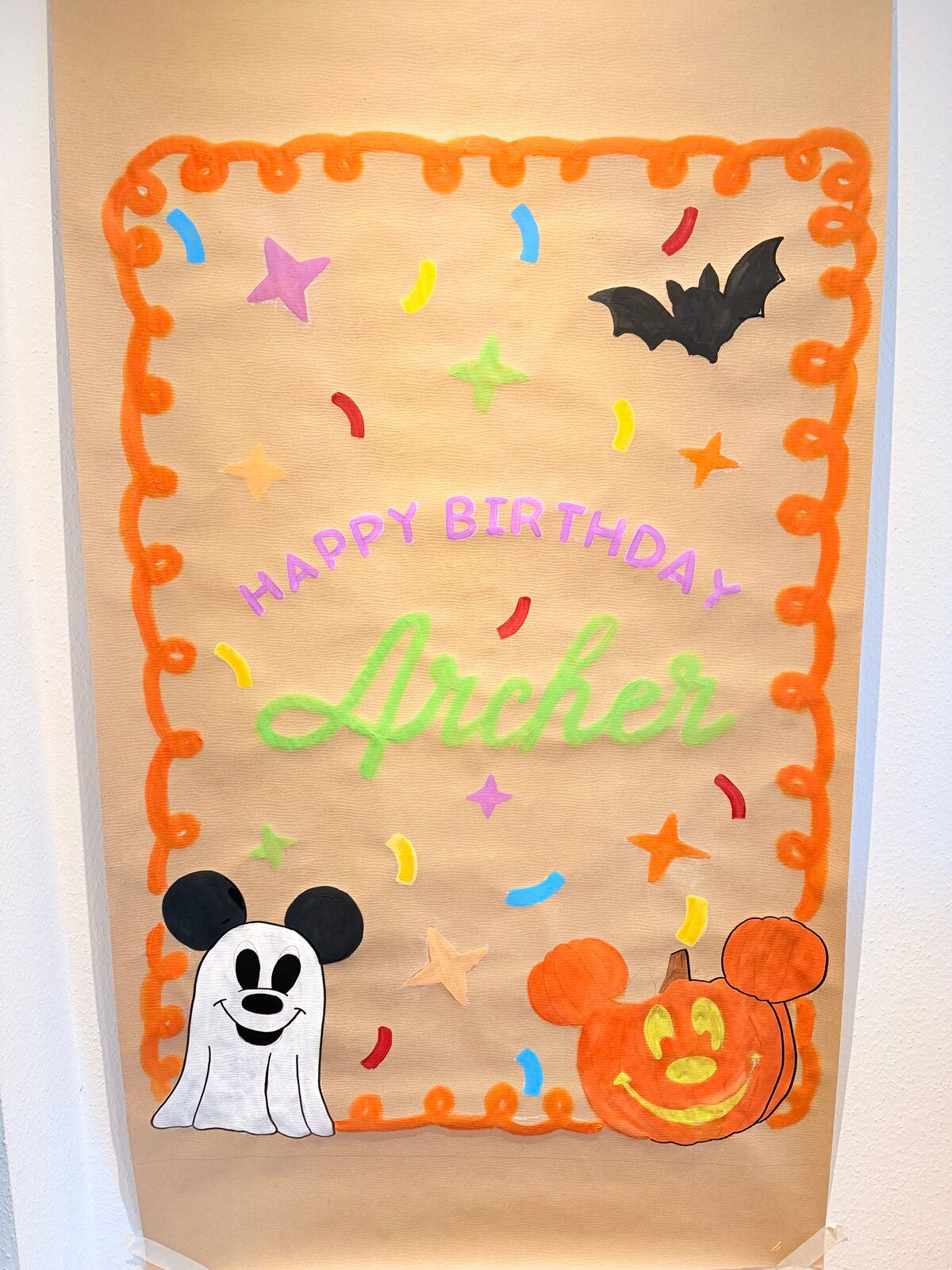 A tall kraft-paper banner hung on a wall, with an orange looped border, confetti and stars. It reads “Happy Birthday Archer”, with a bat, a ghost with mouse ears and a jack-o’-lantern shaped like a mouse head.