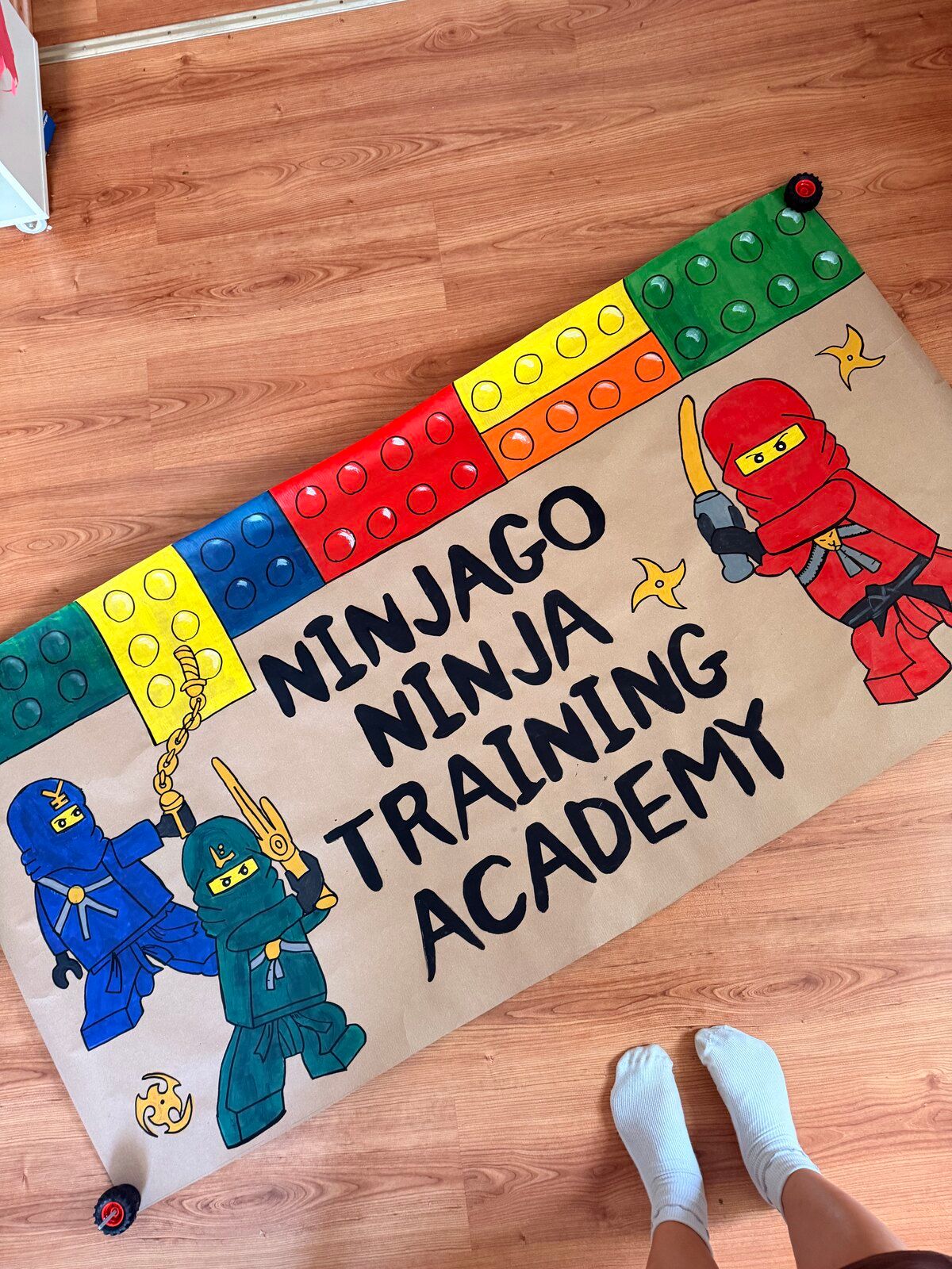 A kraft-paper banner with a row of red, yellow, blue and green toy bricks along the top and three ninja figures in red, blue and green. It reads “Ninjago Ninja Training Academy”.