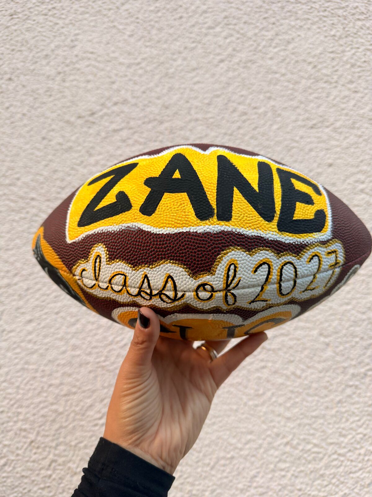 A hand-painted American football in brown and gold, held up against a white wall. It reads “Zane” in black on a gold panel and “class of 2027” in script.