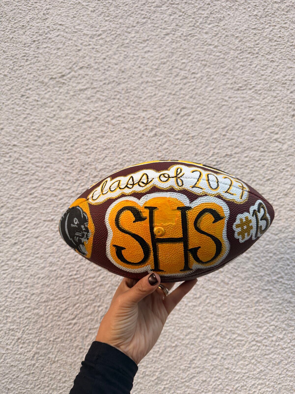 The painted football held up against a white wall, showing “class of 2027”, “SHS” and “#13”.