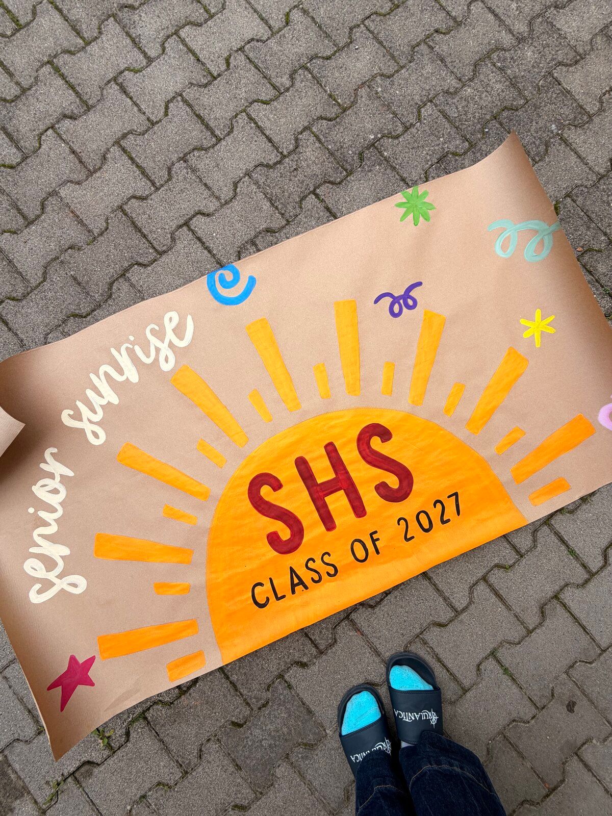 A kraft-paper banner with a big orange half sun and its rays. It reads “senior sunrise” in white script and “SHS Class of 2027” on the sun, with colorful squiggles and stars.