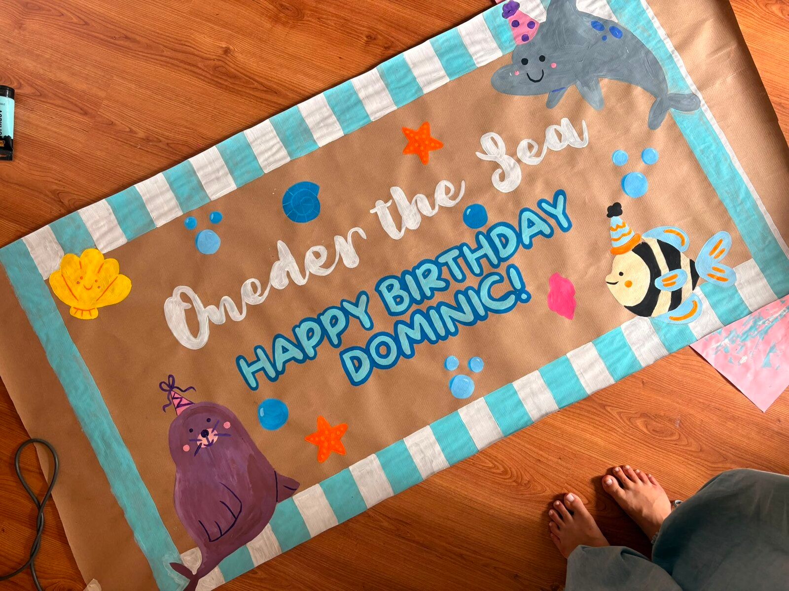 A long kraft-paper banner with a mint-and-white striped border. It reads “Oneder the Sea” in white script and “Happy Birthday Dominic!” in blue, with a dolphin, a seal and a striped fish in party hats, plus shells, starfish and bubbles.